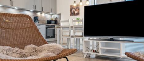 Una sala de estar moderna con un televisor de pantalla plana grande, un sillón de mimbre y un comedor con sillas blancas.