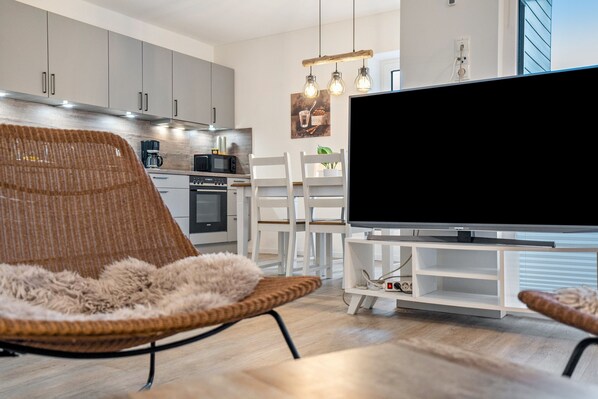 Una sala de estar moderna con un televisor de pantalla plana grande, un sillón de mimbre y un comedor con sillas blancas.