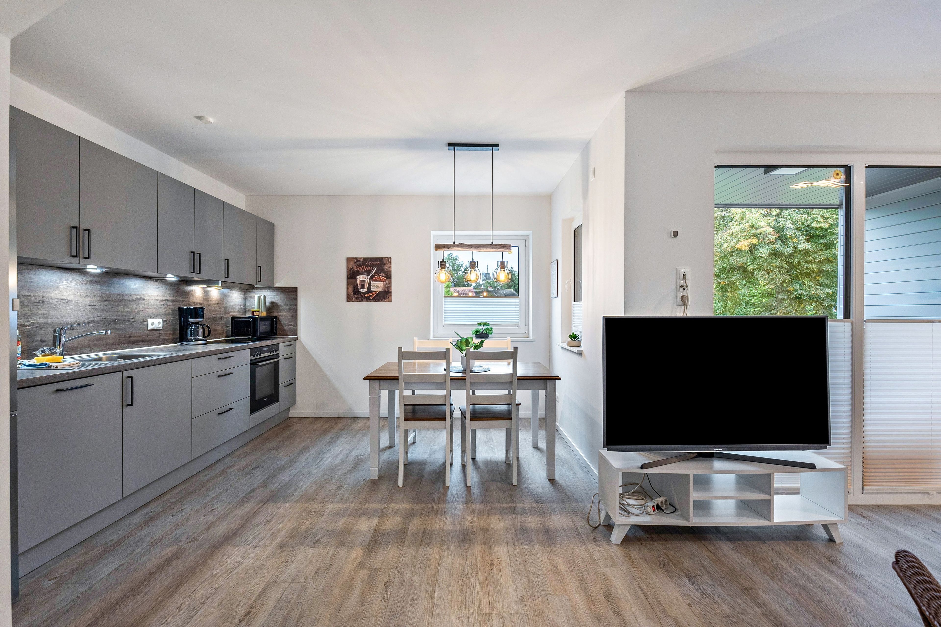 A modern kitchen with grey cabinets, a dining area with a wooden table and chairs, and a flat-screen TV on a white stand.