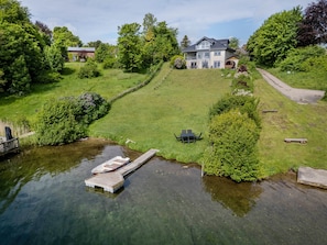 Ett hus med en brygga som sträcker sig ut i ett stilla vattenområde, omgivet av frodig grönska och en sluttande gräsmatta.
