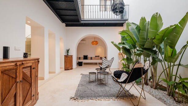 Ein großzügiger Innenbereich mit einer Holzkommode, einer Sitzecke mit Sofa und Sesseln, einer Hängeleuchte und großen Grünpflanzen.