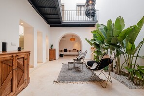 A spacious indoor area with a wooden cabinet, a seating area with a sofa and chairs, a hanging light fixture, and large green plants. - Riad Alaya (علايا) 10mn to The Place - Breakfast (Marrakesh)