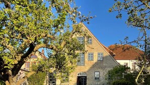 Un edificio de piedra con techo inclinado, un jardín con césped y un árbol de flores rosadas.