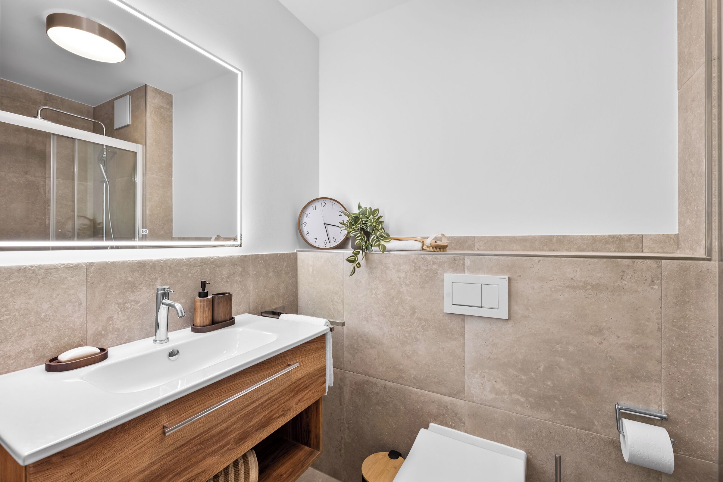 A modern bathroom with a large mirror, a sink with a wooden vanity, a towel rack, a clock, and a small potted plant.