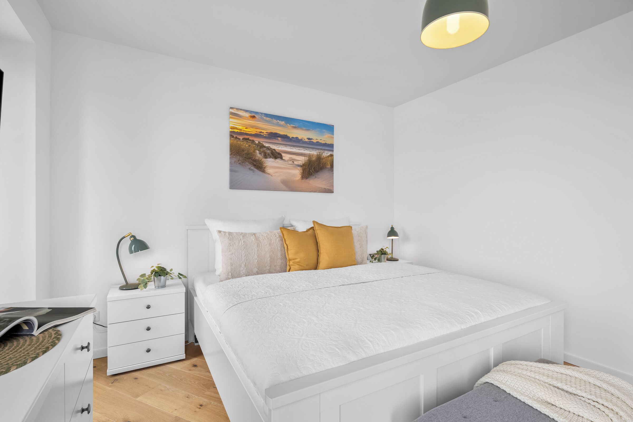 A bedroom with a white bed, a nightstand, a lamp, a plant, and a wall-mounted picture of a beach scene.