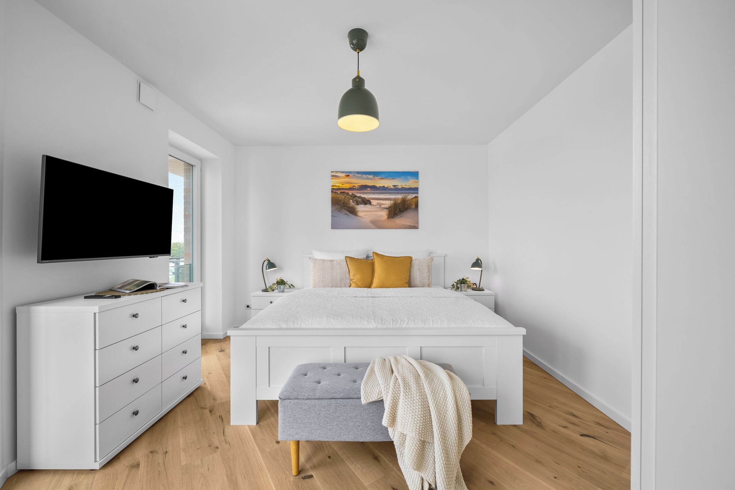 Ein modernes Schlafzimmer mit Flachbildfernseher, einem weißen Bett mit grauem Sitzbank und einem Gemälde mit Strandmotiv, sowie einer Pendelleuchte.