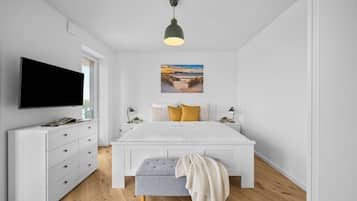 Ein modernes Schlafzimmer mit Flachbildfernseher, einem weißen Bett mit grauem Sitzbank und einem Gemälde mit Strandmotiv, sowie einer Pendelleuchte.