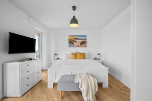 A modern bedroom with a flat-screen TV, a white bed with a grey bench, a painting of a beach scene, and a pendant light. - The View A08 Belvedere Nordsee - Balcony,sea view,garden furniture,shared. Sauna (Dagebüll)