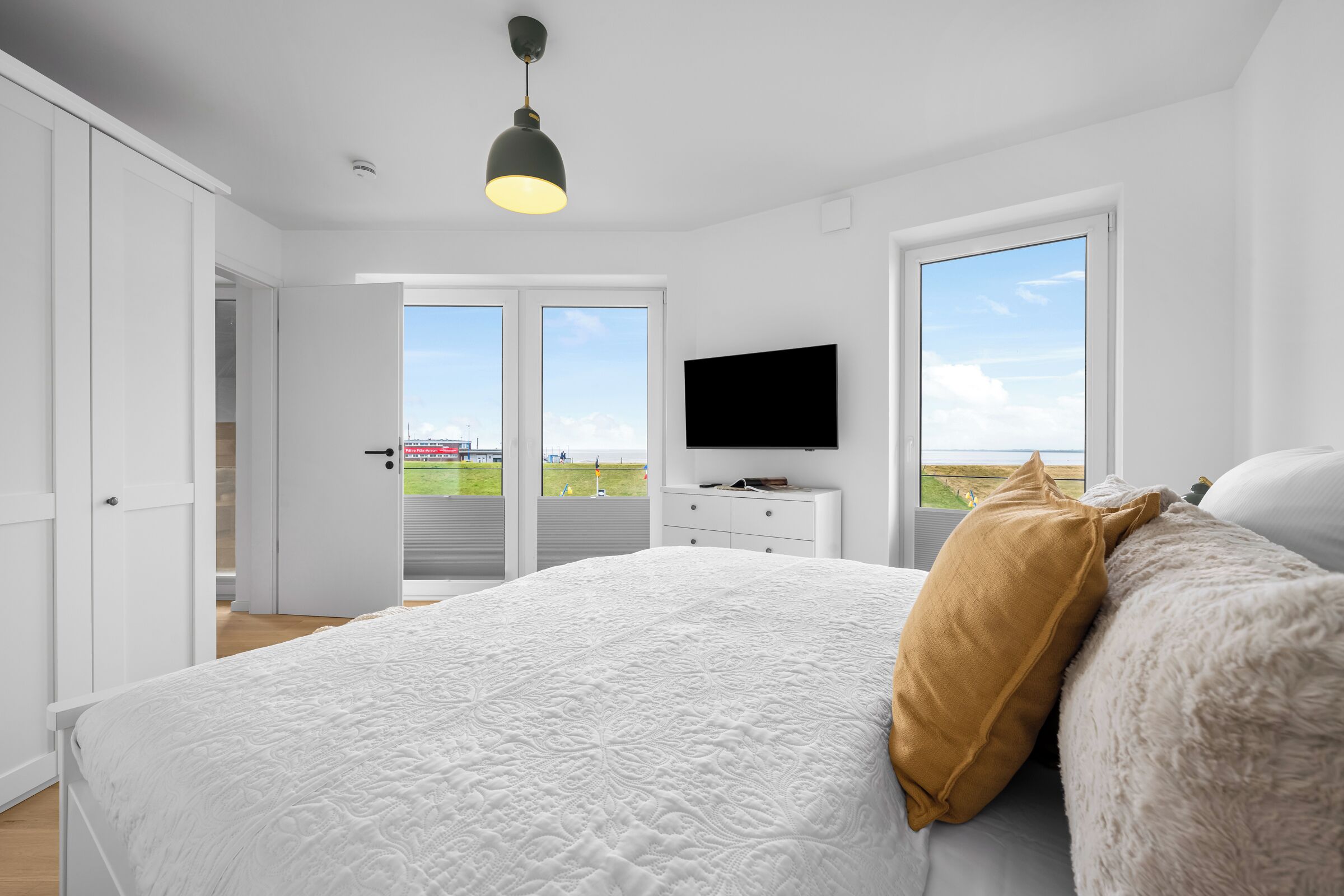 A bedroom with a large bed, a television on a dresser, and a view of the beach through the windows.