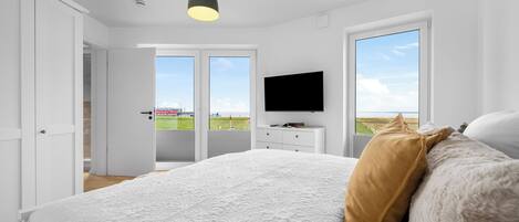 A bedroom with a large bed, a television on a dresser, and a view of the beach through the windows.