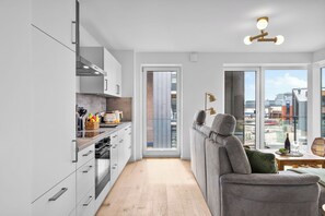A modern kitchen with white cabinets, a dining area with a grey sofa, and a view of the city through large windows. - The View A08 Belvedere Nordsee - Balcony,sea view,garden furniture,shared. Sauna (Dagebüll)