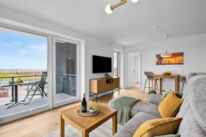 A modern living room with a large sliding glass door leading to an outdoor area, a flat-screen TV mounted on the wall, and a dining area with a table and chairs. - The View A08 Belvedere Nordsee - Balcony,sea view,garden furniture,shared. Sauna (Dagebüll)