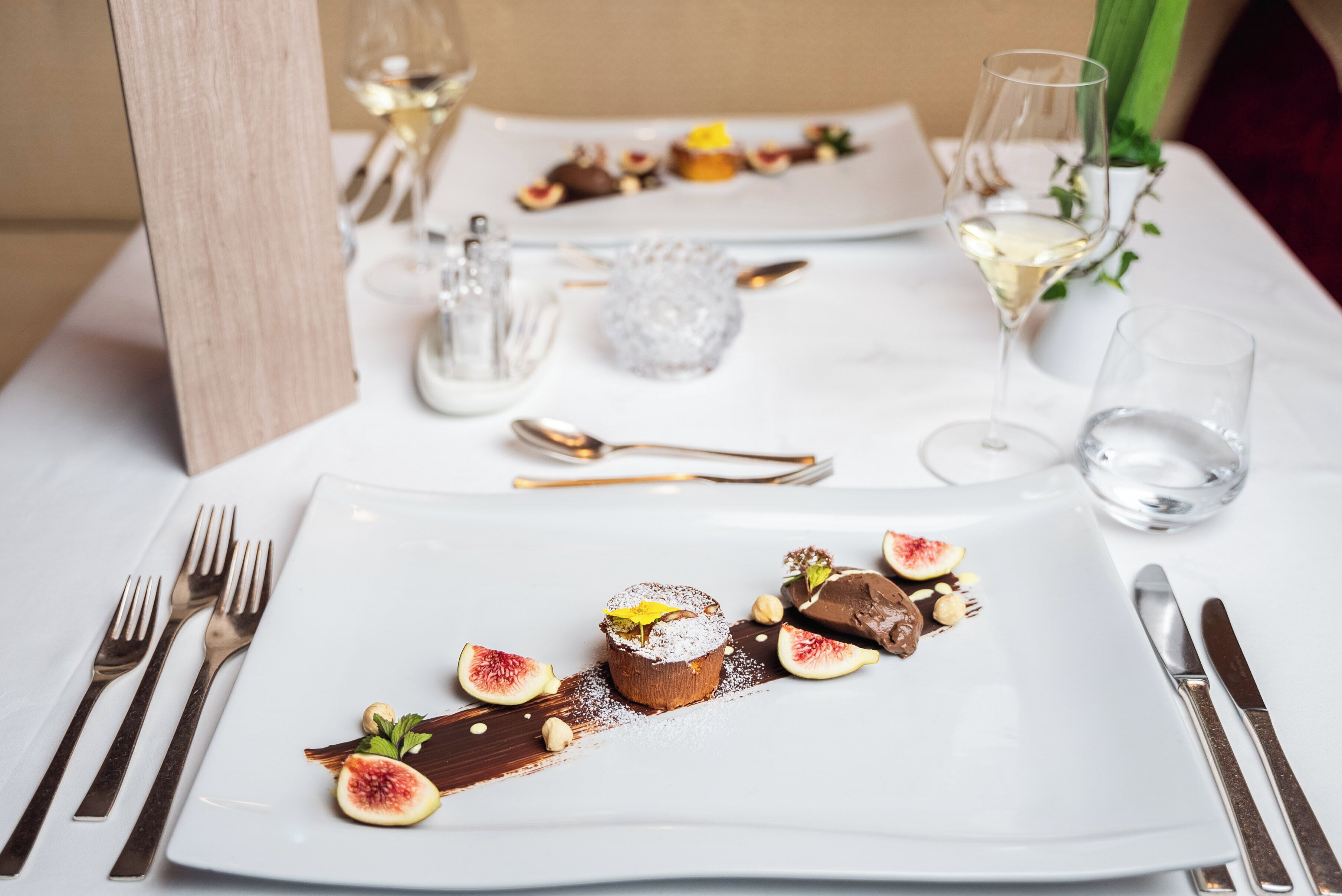 A dining table with a dessert plate, cutlery, and a glass of white wine.