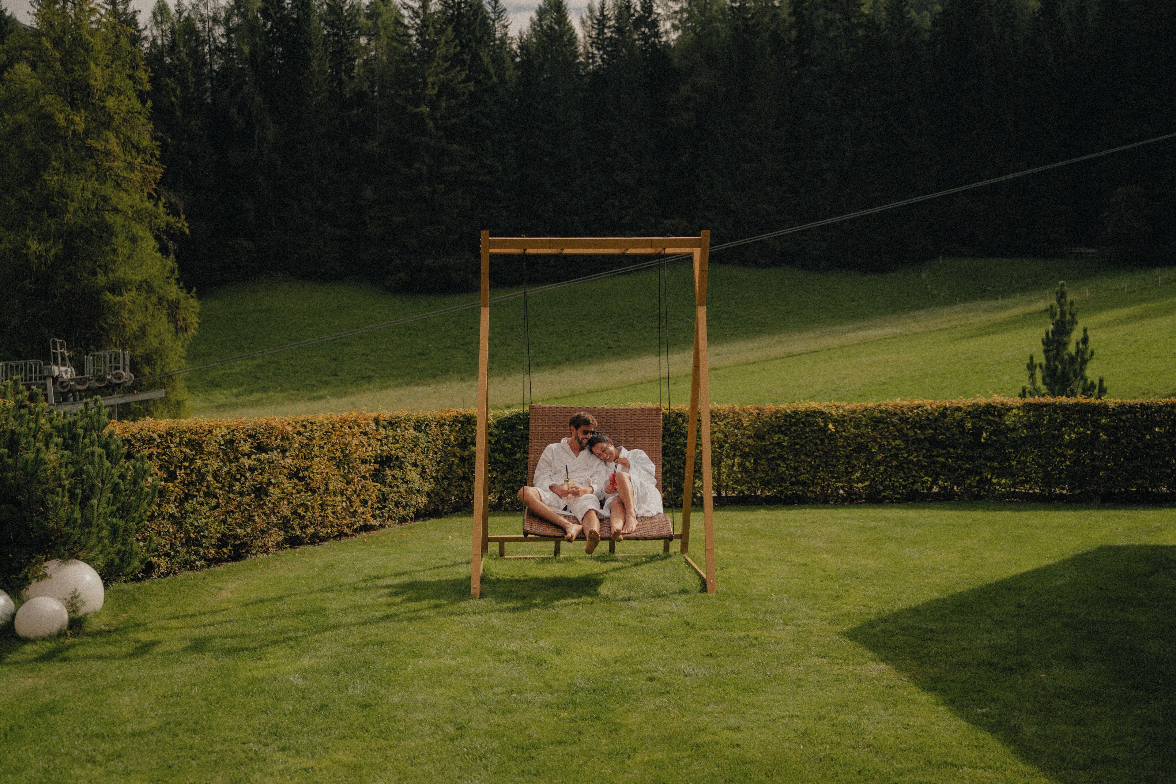 A couple sitting on a swing in a grassy area with a hedge and trees in the background.