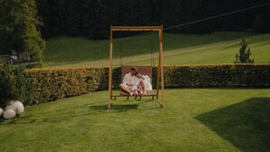 Ein Paar sitzt auf einer Schaukel in einer grünen Anlage mit Hecke und Bäumen im Hintergrund.