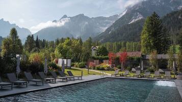 A pool area with lounge chairs and a small building, surrounded by trees and mountains.