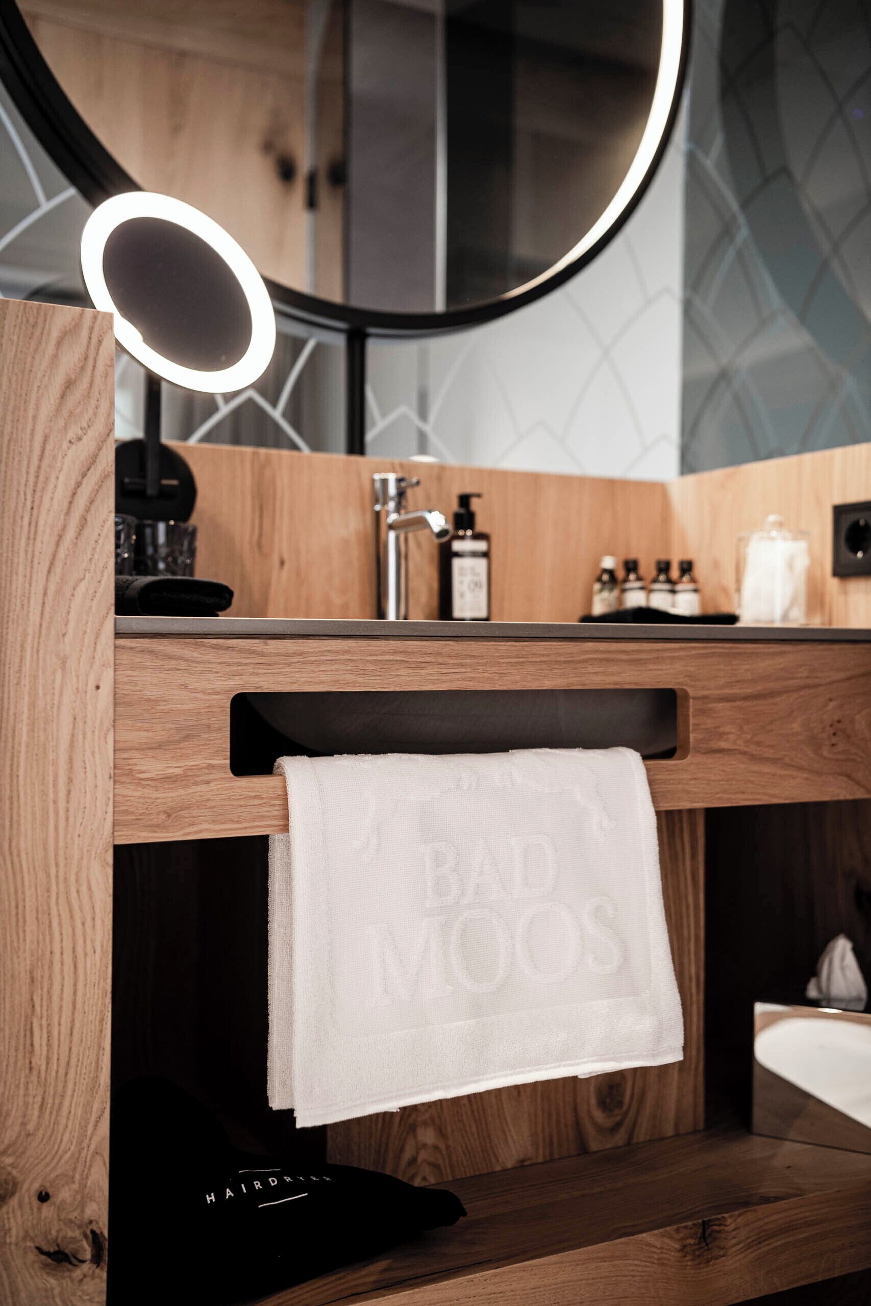 A bathroom with a wooden vanity, a mirror with a light, a sink, and a towel hanging on a rack.