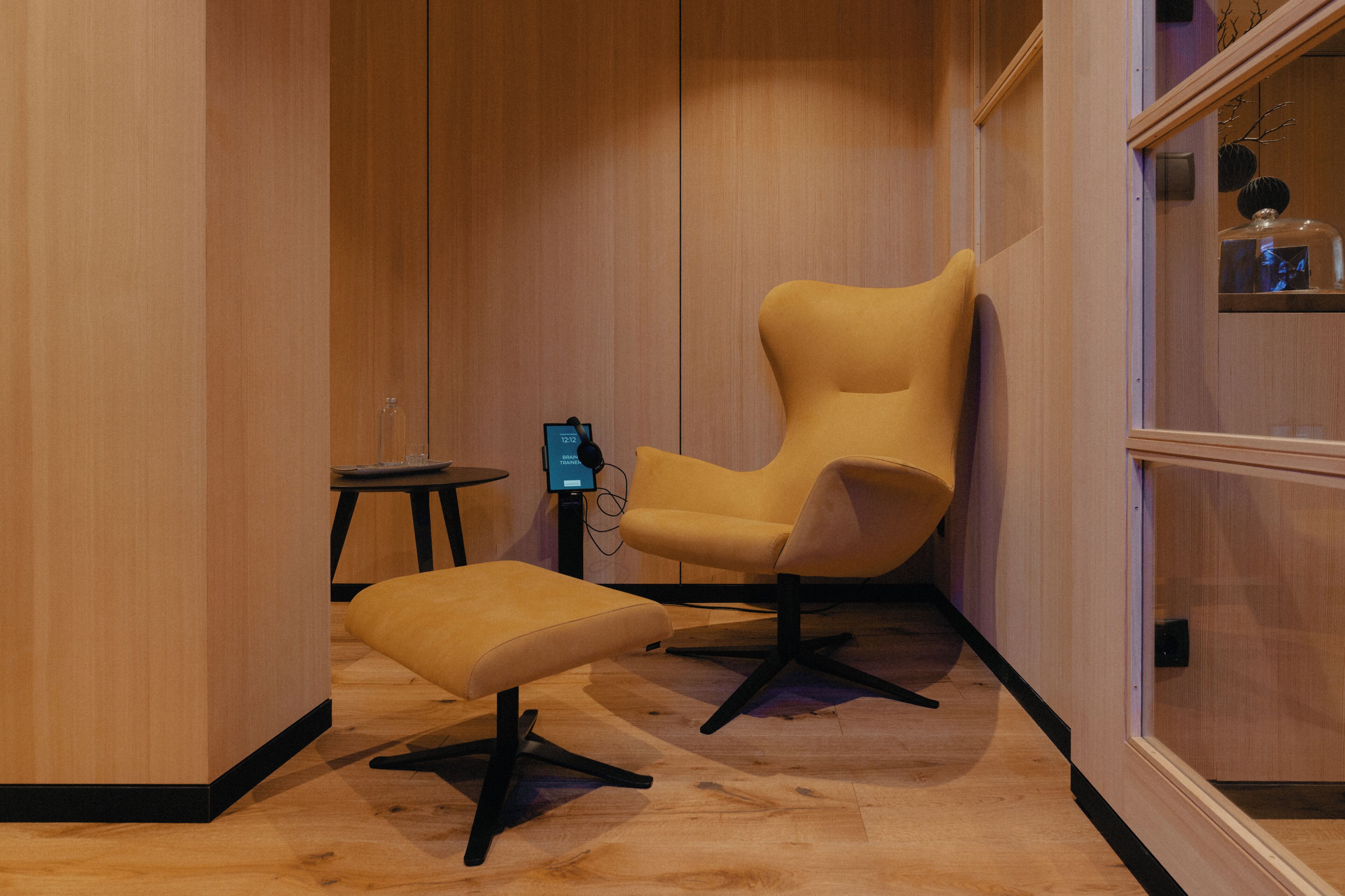 Une chambre moderne avec un plancher en bois, un fauteuil jaune, un pouf assorti et une petite table sur laquelle se trouve un appareil numérique.