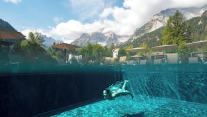 Ein Schwimmbad mit einer Person, die schwimmt, umgeben von Bergen und Außenbereich mit Sitzgelegenheiten.