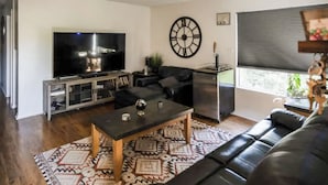A living room with a flat-screen TV, a large wall clock, a sofa, a coffee table, and a patterned rug. - Escondido Urban Stay - 2BR Apartment (Escondido)