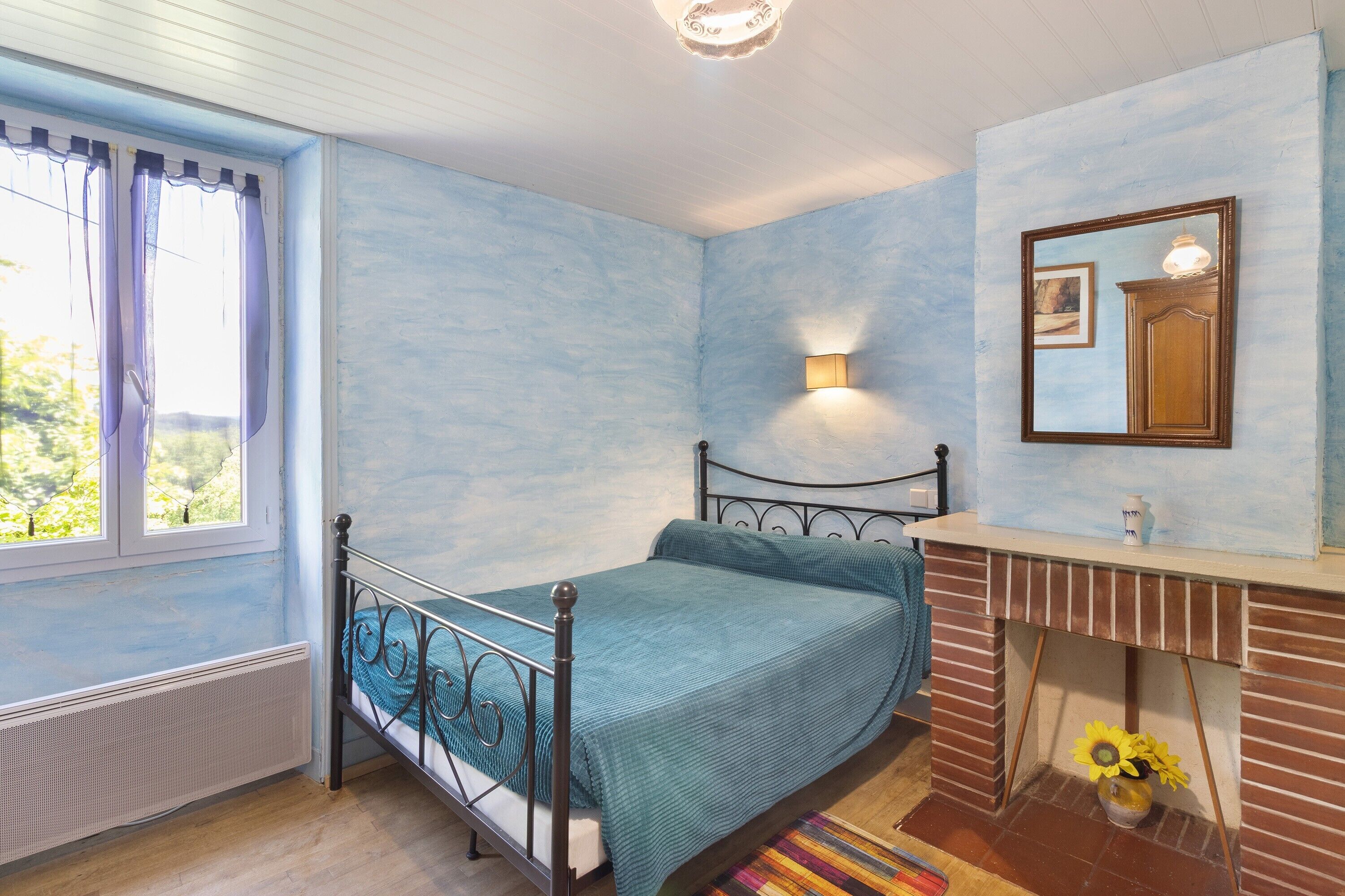A bedroom with a bed, a radiator, a window with curtains, a wall-mounted light, and a mirror.