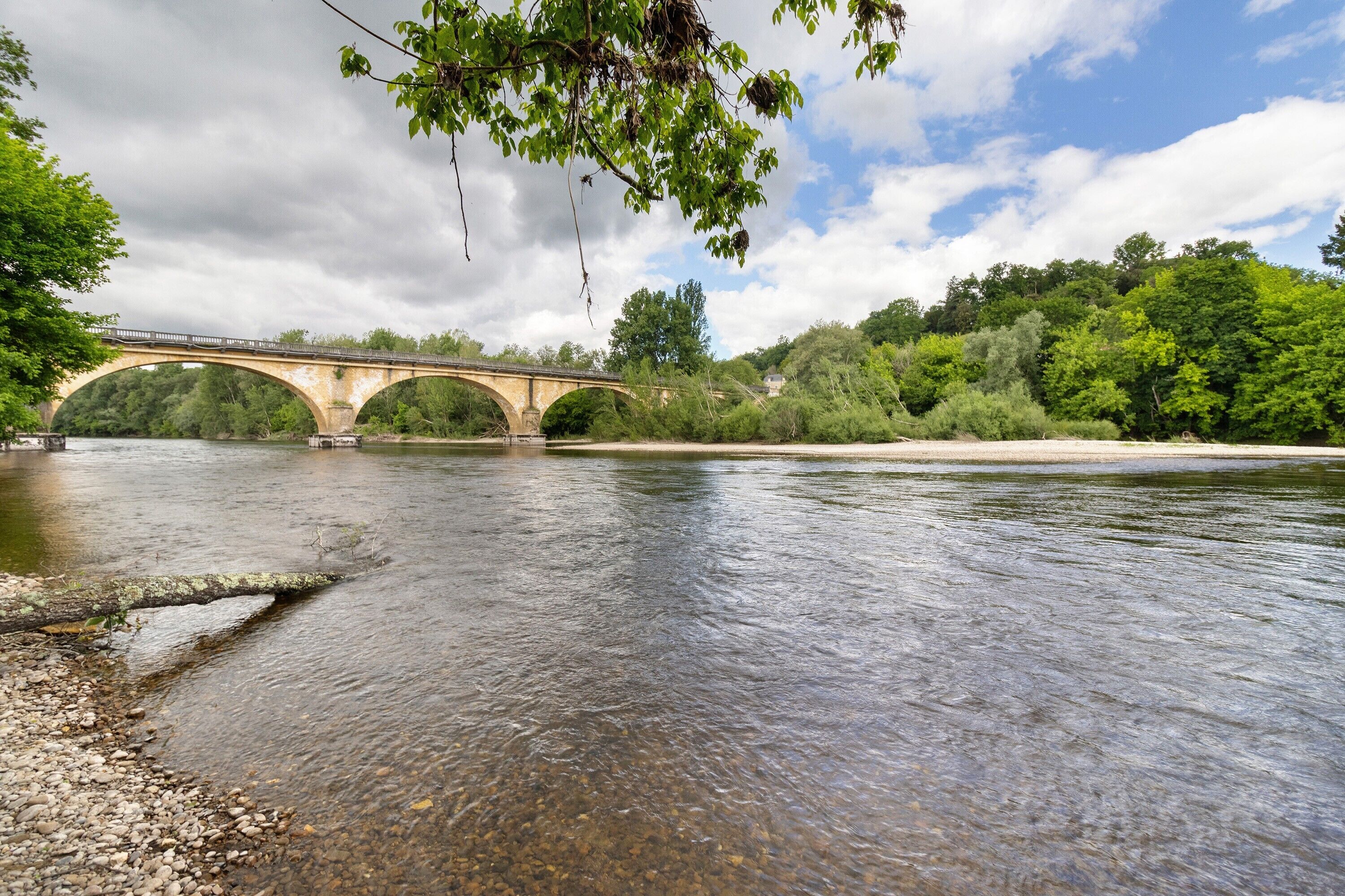 Een stenen brug over een rivier met helder water, omringd door bomen en begroeiing.