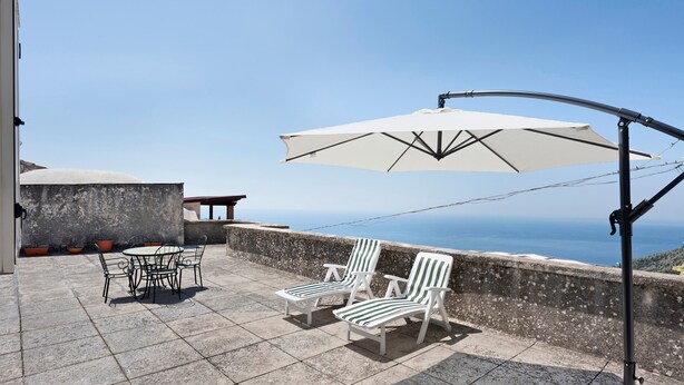 Um pátio com mesa e cadeiras, um guarda-sol branco e vista para o mar.