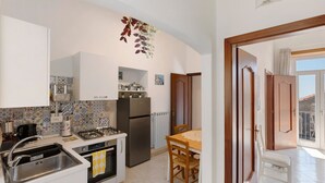 Cozinha compacta com pia, fogão e geladeira, mesa de jantar com cadeiras, e vista para o exterior através de uma porta.