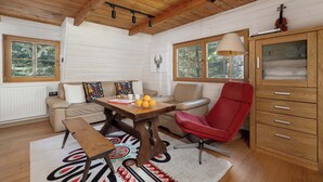 Ein gemütliches Blockhaus-Interieur mit Holzbalkendecke, einem Esstisch mit einer Bank, einem roten Stuhl, einem Schrank und großen Fenstern.