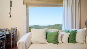 A room with a sofa, a dining table with chairs, and a window with a view of green hills. - Apartment 'Agriturismo Il Ghiaccio - Mirto' with Sea View, Wi-Fi and Air Conditioning (Suvereto)