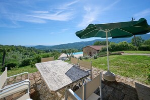 Eine Terrasse im Freien mit Tisch und Stühlen, einem grünen Sonnenschirm und Blick auf Berge und ein Schwimmbad.