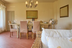 A dining area with a table set for four, chairs, a chandelier, a painting, a mirror, and a wicker bench. - Apartment 'Agriturismo Il Ghiaccio - Oleandro' with Sea View, Wi-Fi and Air Conditioning (Suvereto)