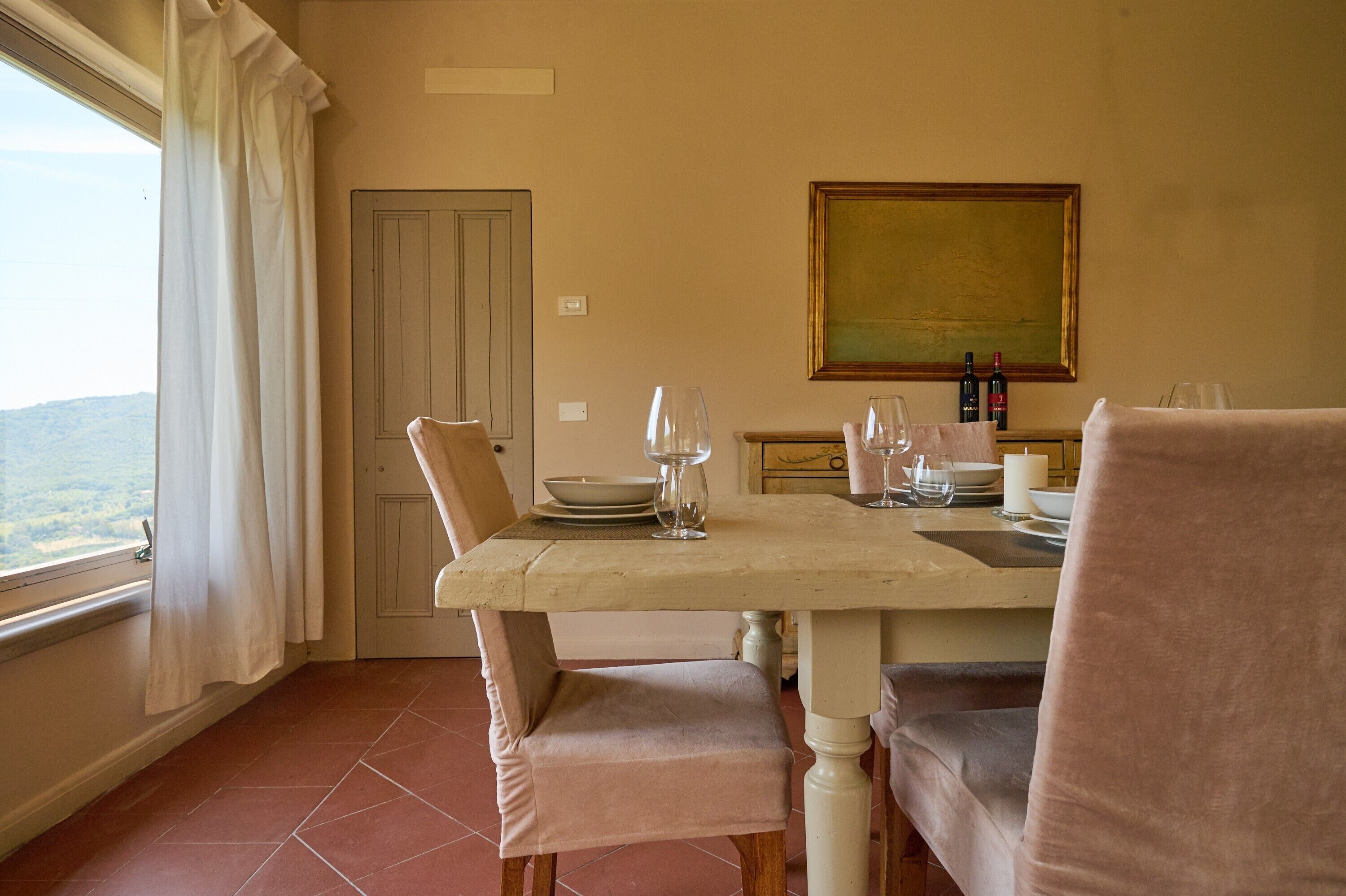 A dining area with a table set for two, a painting on the wall, and a view through the window.
