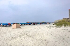 A beach with umbrellas and lounge chairs, a pier in the background, and a building on the right.