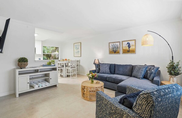 A modern living room with a grey sofa, a round coffee table, a white entertainment unit, and a dining area with a white table and chairs. - Under 1 Mile to Siesta Key Beach | Beach Gear Included | Pool | Sleeps 4 (Sarasota)