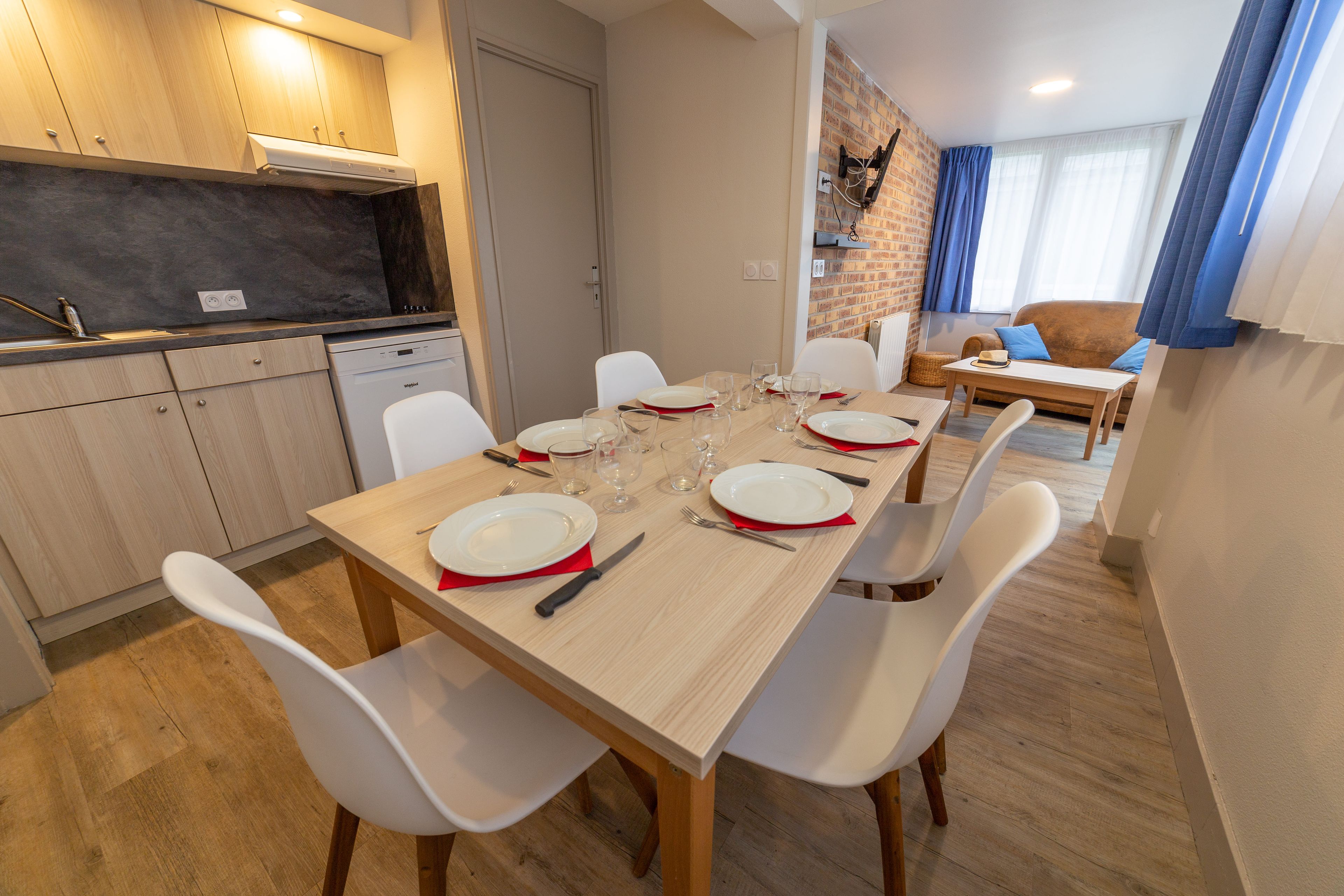 A dining area with a wooden table set for six, white chairs, and a kitchen in the background.
