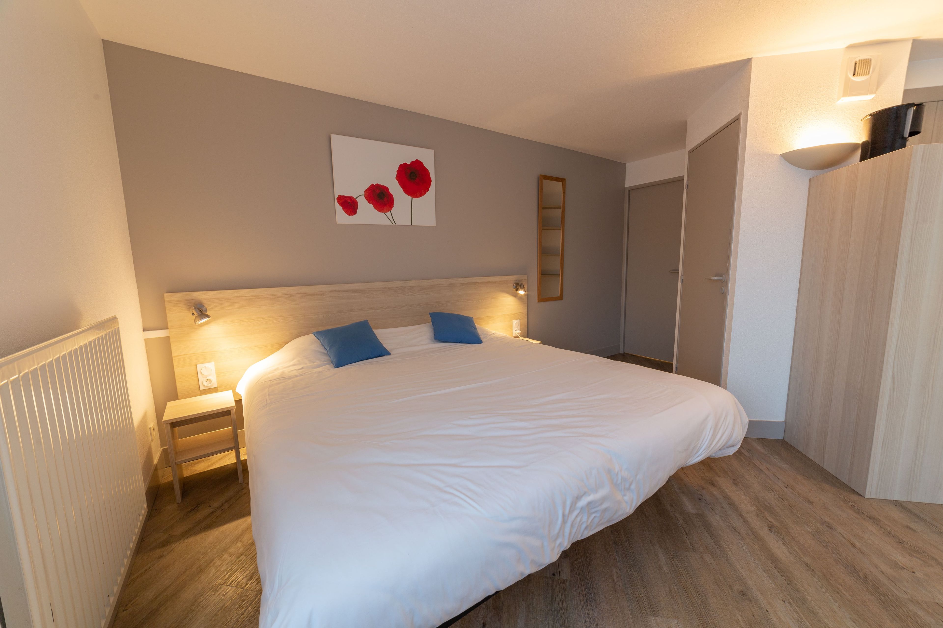 Un lit bien fait, avec une literie blanche et des oreillers bleus, une table de chevet avec une lampe et un miroir fixé au mur.