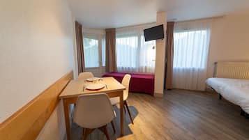 A hotel room with a wooden table set for two, a bed, a TV mounted on the wall, and a window with curtains.