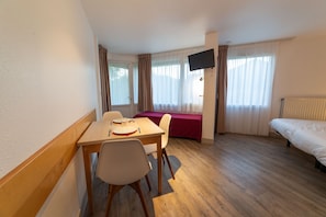 A hotel room with a wooden table set for two, a bed, a TV mounted on the wall, and a window with curtains.
