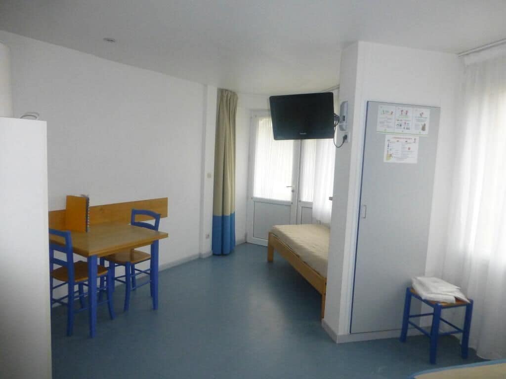 A single room with a bed, a desk with chairs, a television mounted on the wall, and a small stool.