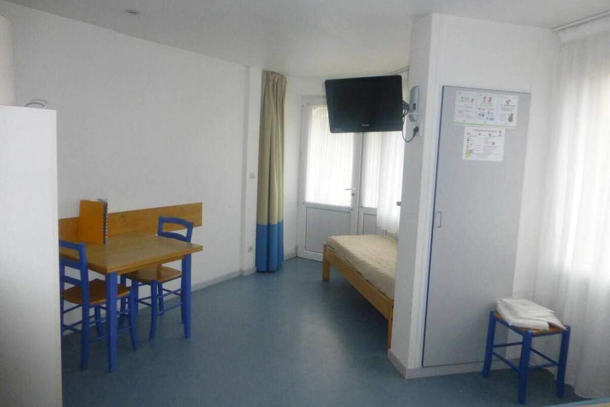 Una habitación individual con una cama, un escritorio con sillas, un televisor montado en la pared y un taburete pequeño.