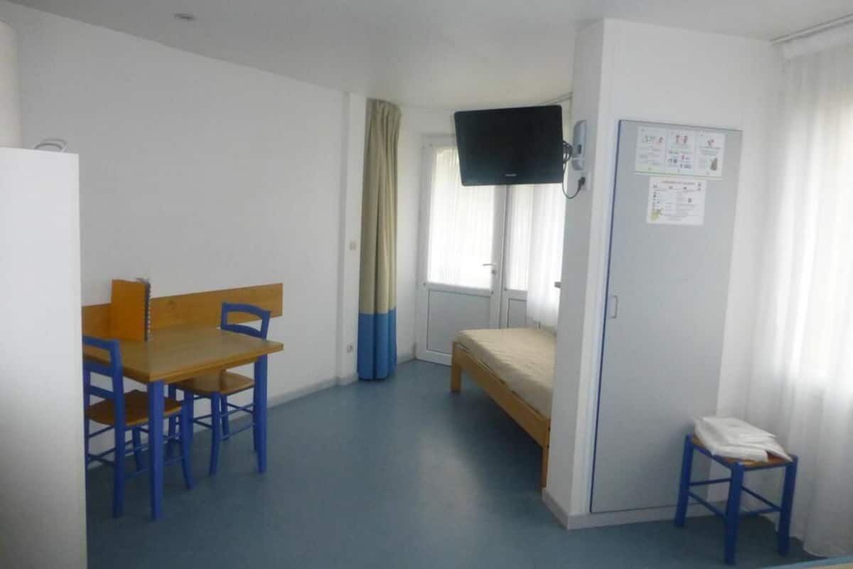 Une chambre individuelle comprenant un lit, un bureau avec des chaises, un téléviseur fixé au mur et un petit tabouret.