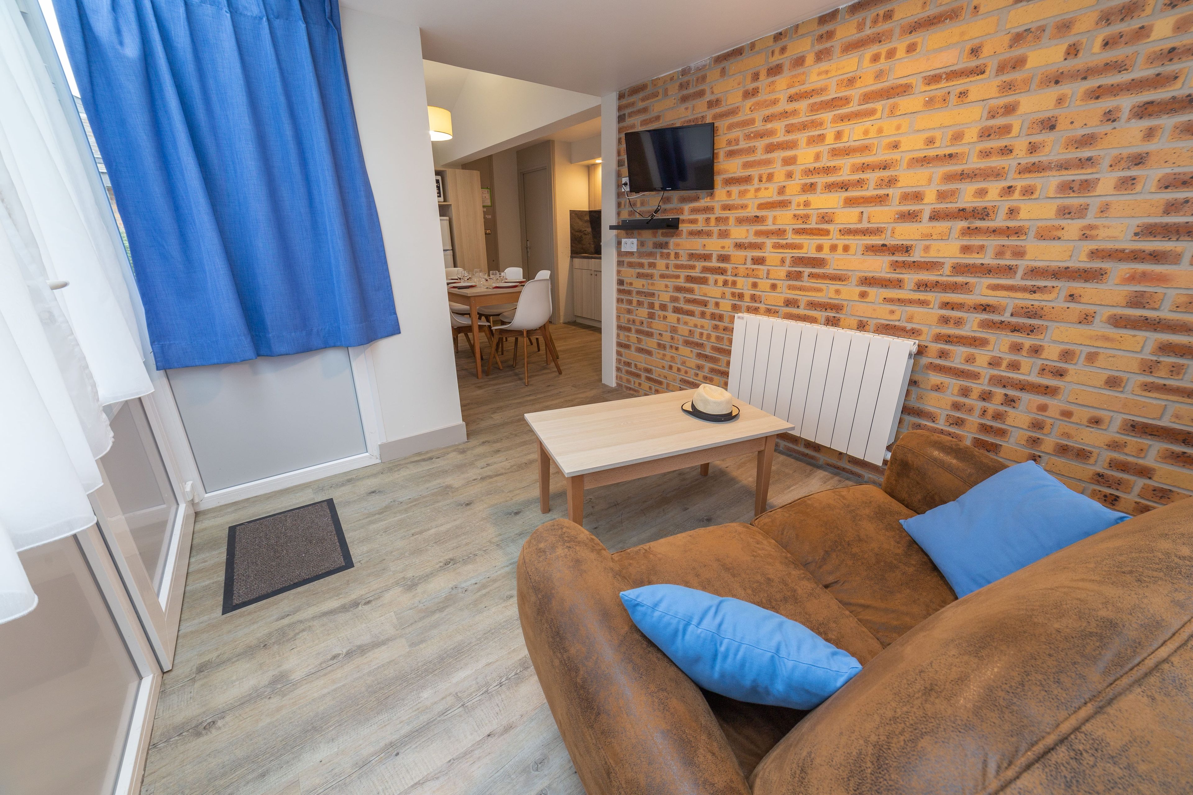 Una sala de estar moderna con una pared de ladrillo, una mesa de madera, un sofá con cojines azules y un televisor montado en la pared.
