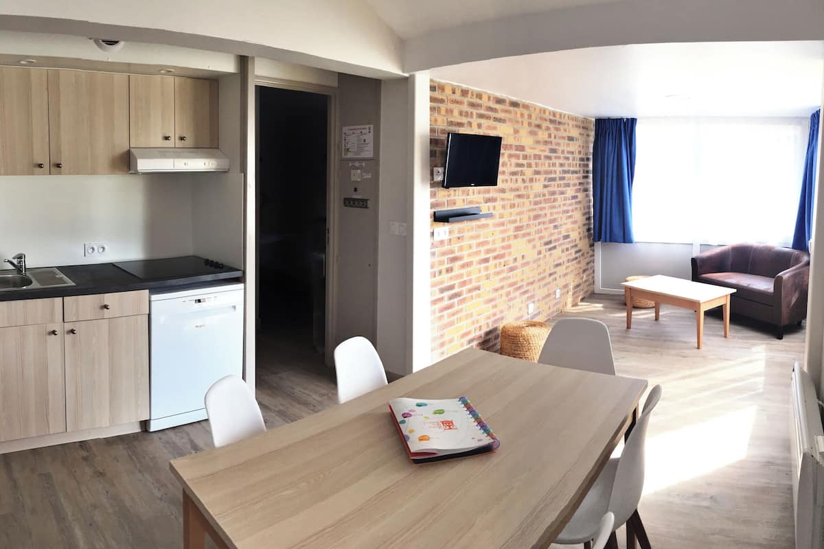 Un espacio habitable compacto con kitchenette, comedor, zona de estar con televisor y pared de ladrillo.