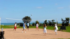 Eine Gruppe spielt Beachvolleyball auf einem Sandplatz, umgeben von Grünflächen und einem klaren Himmel.