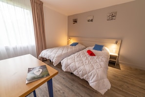 A hotel room with two beds, a wooden table, and a window with curtains.
