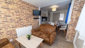 A compact living space with a brick wall, a sofa, a dining table with chairs, and a television mounted on the wall.