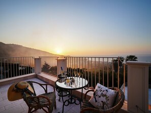 Ein Balkon mit Korbmöbeln, einem Glastisch und Blick auf den Sonnenuntergang über einer Stadtlandschaft.