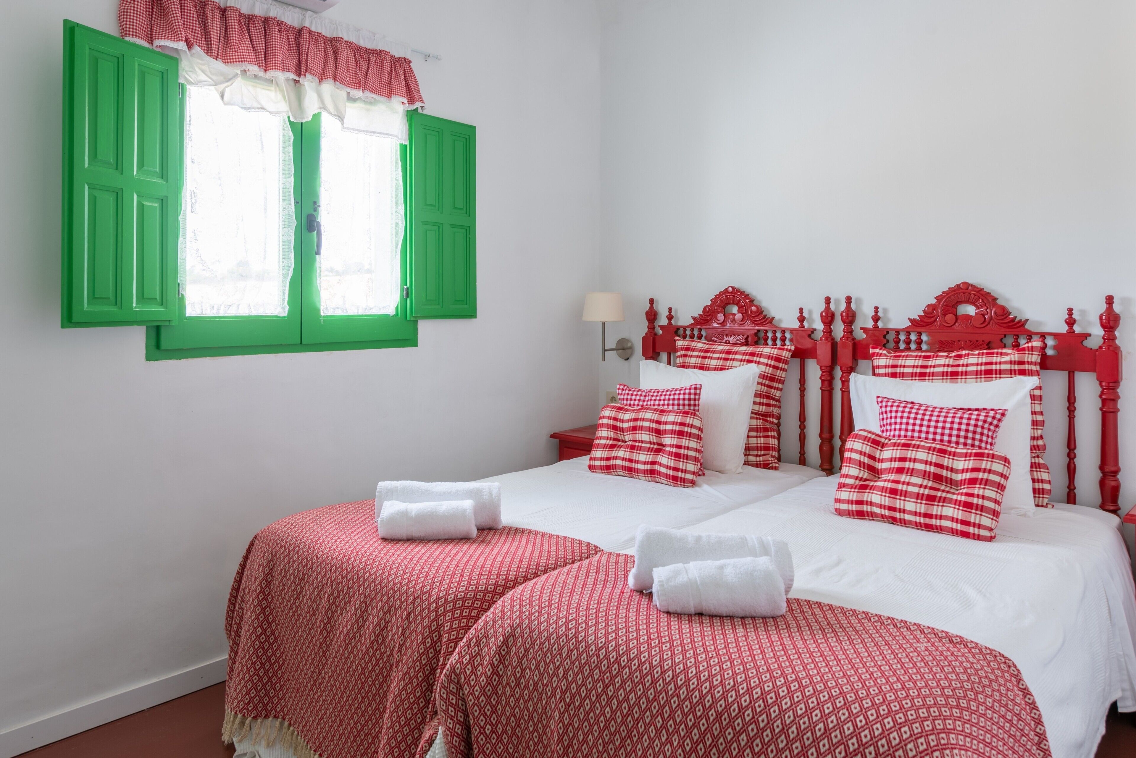 Un lit double avec une literie rouge et blanche, une tête de lit rouge et une fenêtre avec des volets verts.