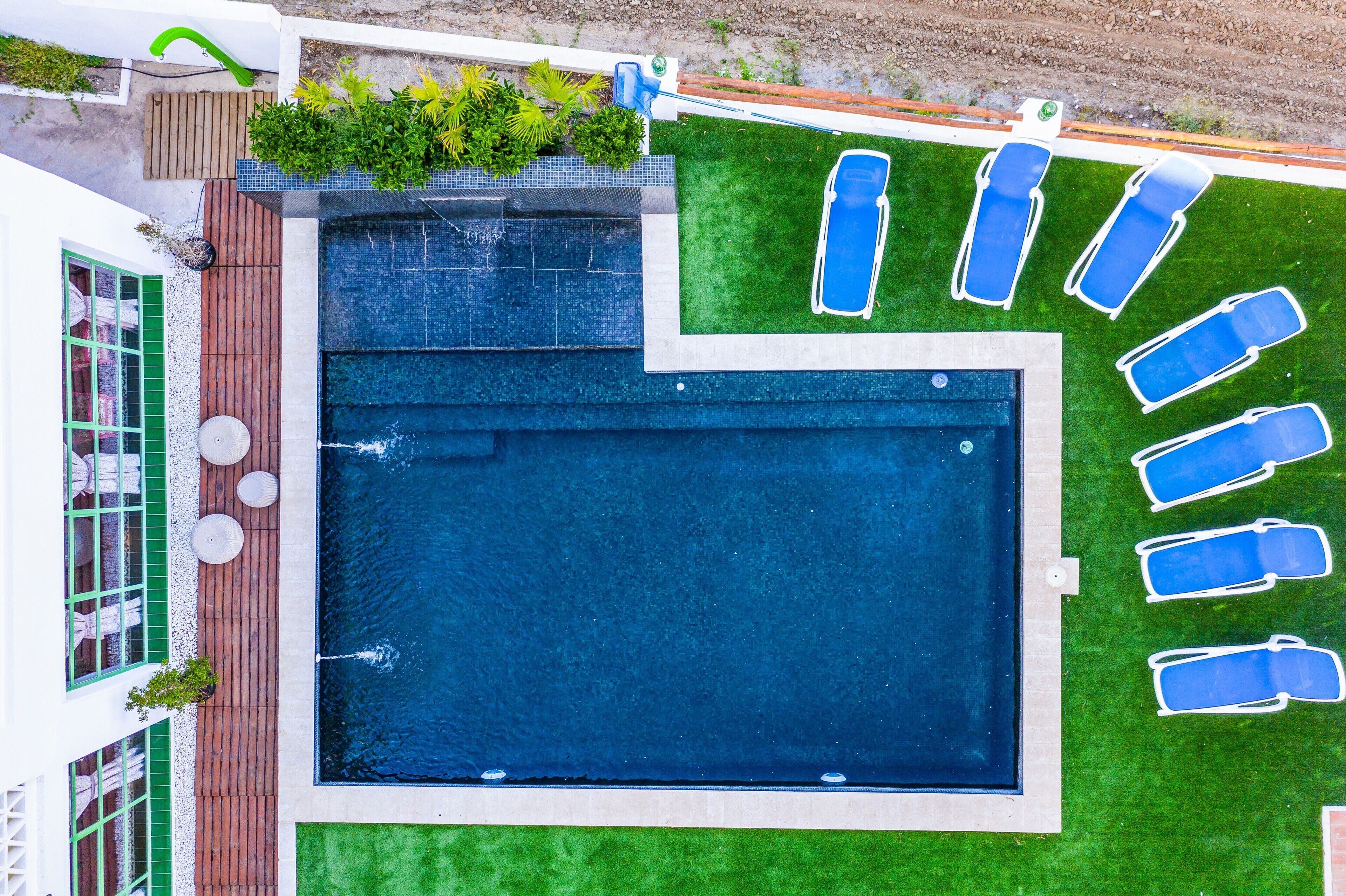 Una piscina rectangular con una solana circundante, un pequeño jardín con plantas y una fila de sillas reclinables azules.